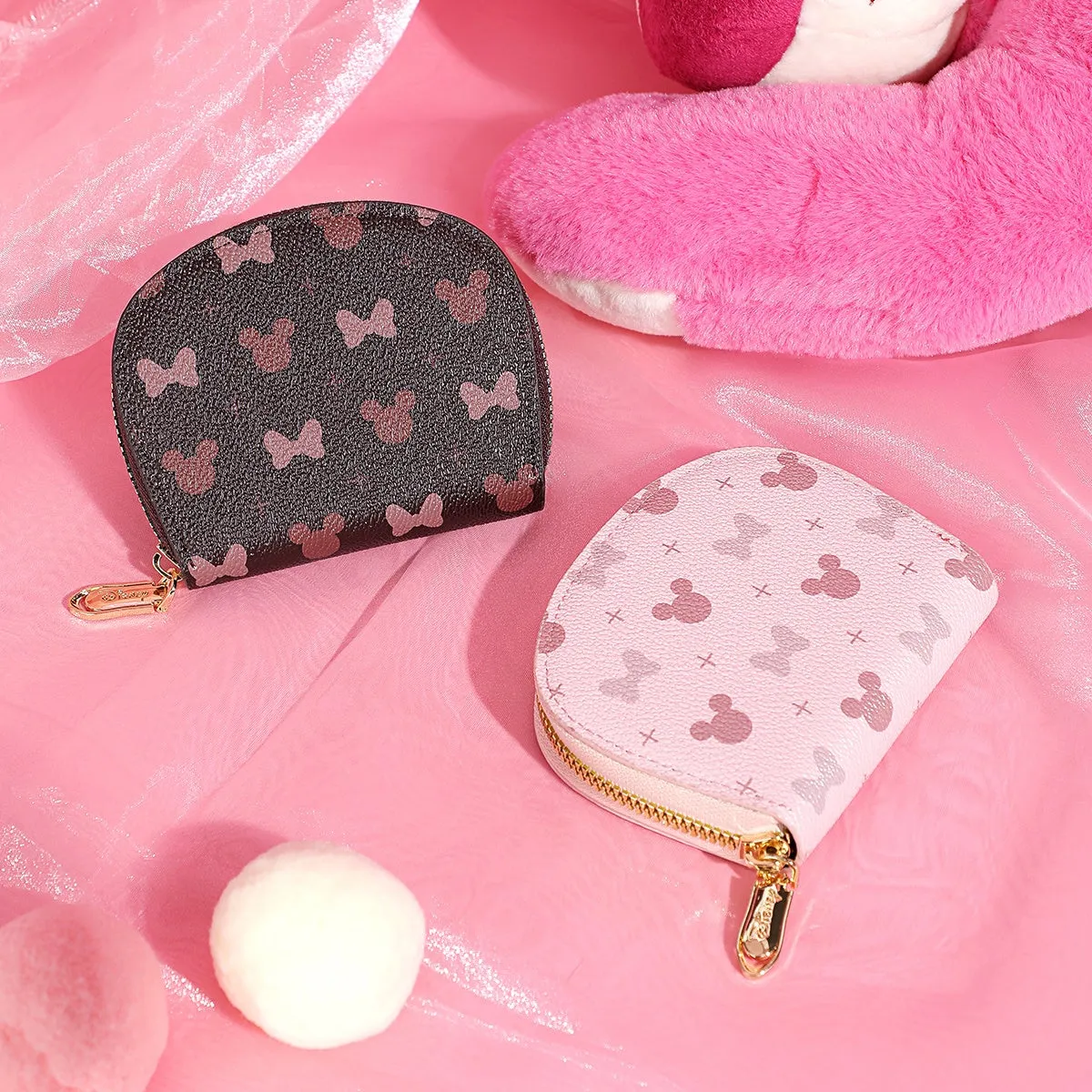 ピンク色の背景に、ミニーマウスのモチーフが描かれた2つの財布が置かれています。黒い財布はミニーマウスのモチーフが全面に、ピンクの財布はミニーマウスとクロス模様が描かれています。背景には、ピンク色のぬいぐるみと白いポンポンも配置されています。