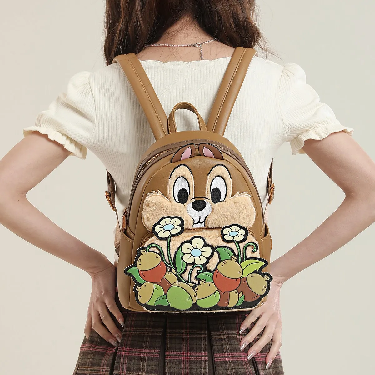 女性が背負っている茶色のリュックサック。リュックサックにはディズニーのキャラクター、チップとデールが描かれており、花とドングリの装飾が施されている。女性は白いシャツとチェックのスカートを着用しており、背景は白。
