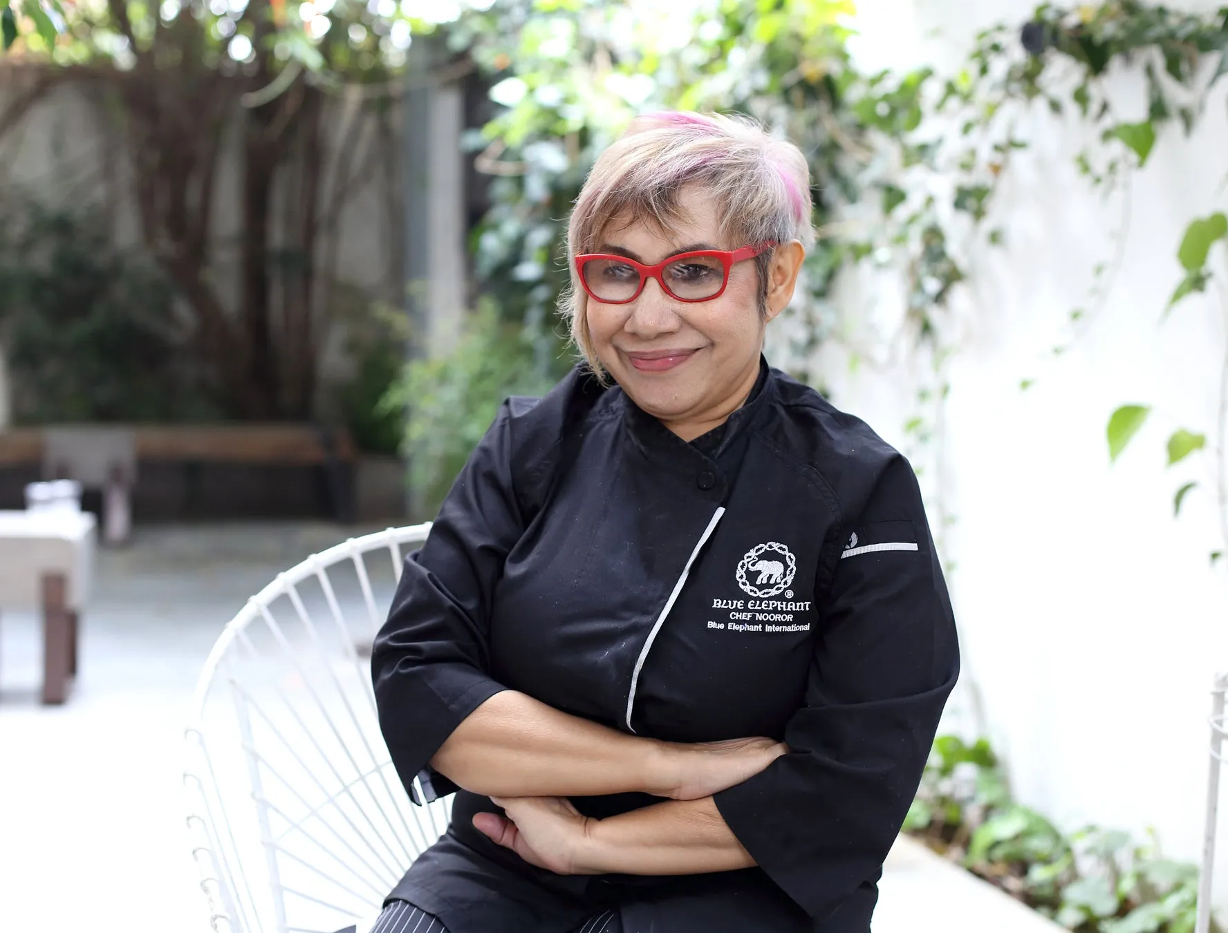 ピンクの髪の女性が、赤いメガネをかけ、黒いシェフコートを着て、腕を組んで座っています。コートには「Blue Elephant」のロゴが刺繍されています。