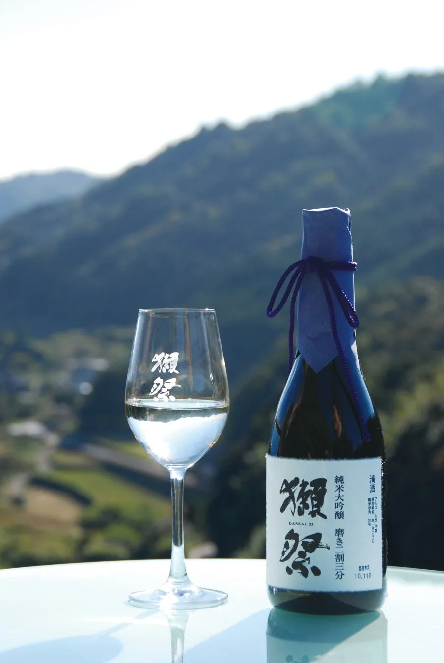 日本酒の獺祭23とグラスが置かれた写真。背景には山々が写っています。