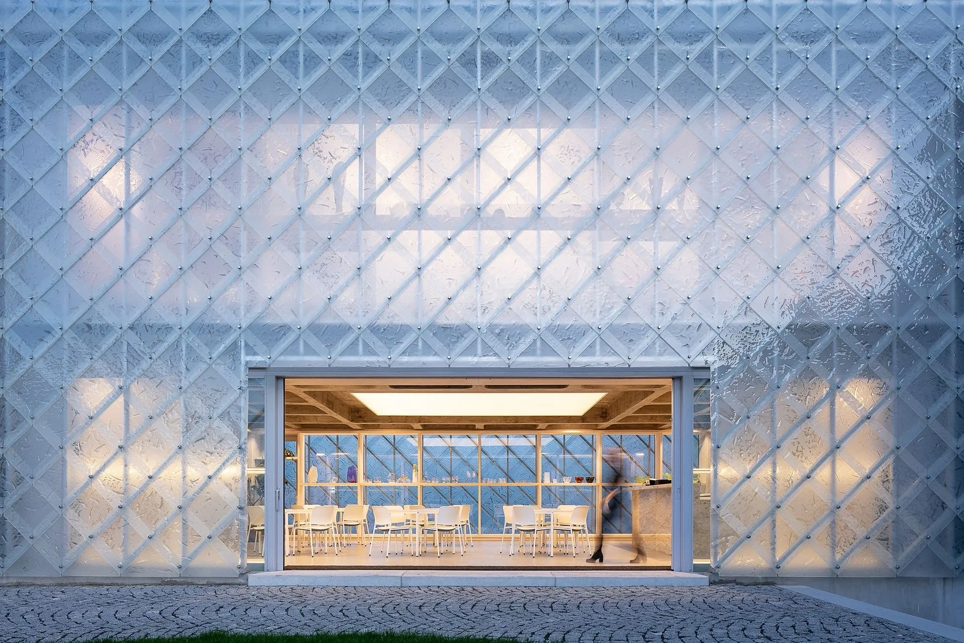 透明なガラス張りの壁を持つ建物の外観。建物の中には、テーブルと椅子が配置され、人が歩いている様子が見える。建物の外は石畳になっている。
