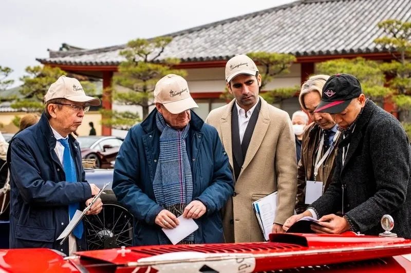 クラシックカーイベントで集まる人々。男性たちは、赤いクラシックカーを囲んで書類を見ています。背景には、日本の寺院の建物が見えます。