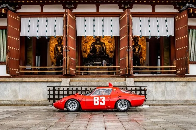 お寺の前に赤いレーシングカーが停まっている写真です。車体には「83」の文字が白く書かれています。お寺の入り口には仏像が安置されており、日本の伝統的な風景とモータースポーツが融合した印象的な一枚です。