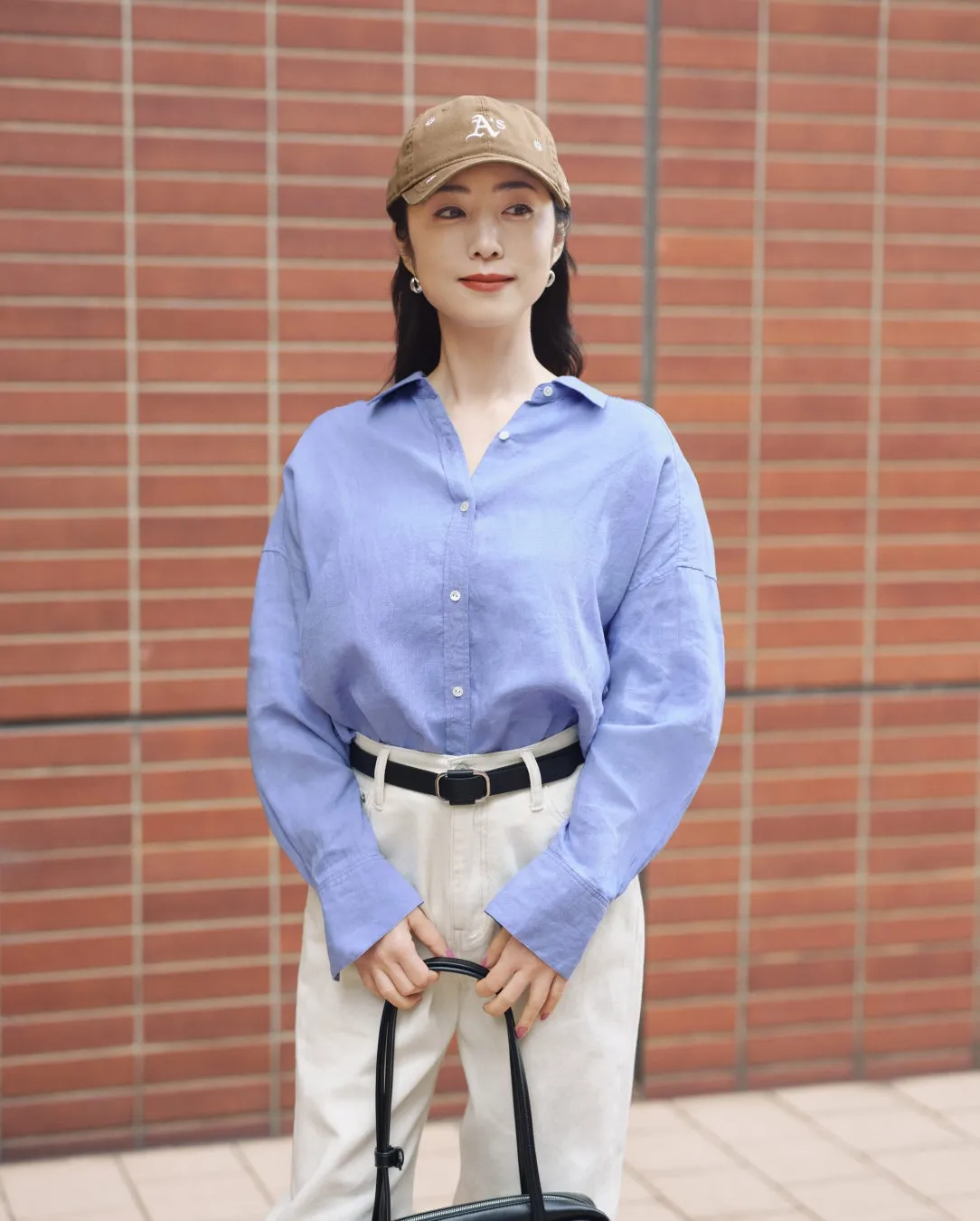 女性が帽子とシャツを着て、バッグを持っています。レンガの壁を背景に立っています。