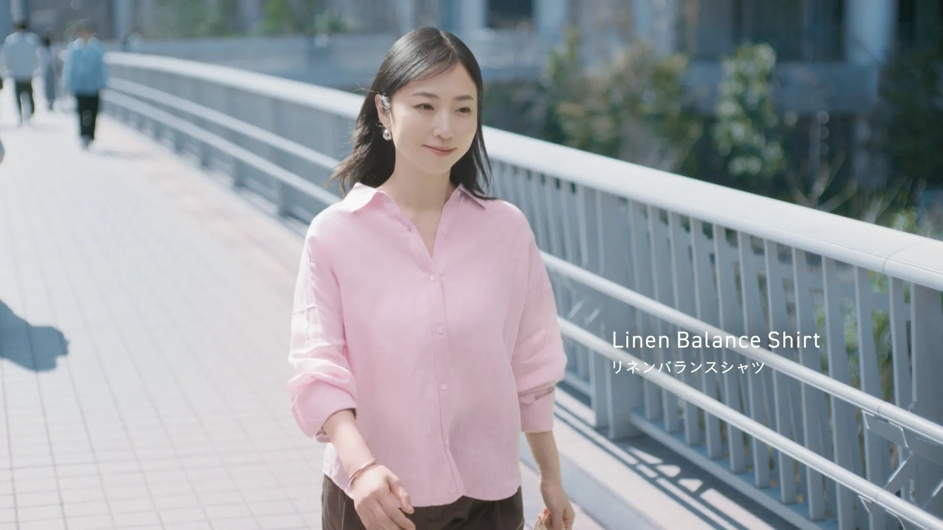 ピンク色のリネンシャツを着た女性が屋外の橋を歩いている様子。