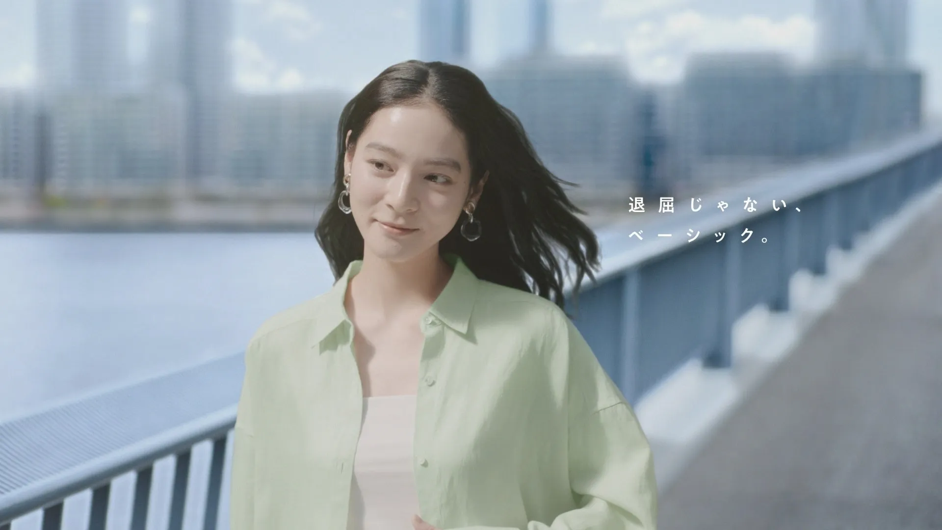 女性が橋を歩いている様子を捉えた写真。彼女はライトグリーンのシャツと白いインナーを着ており、笑顔でカメラを見つめている。背景には水辺と都市の建物がぼやけて写っている。