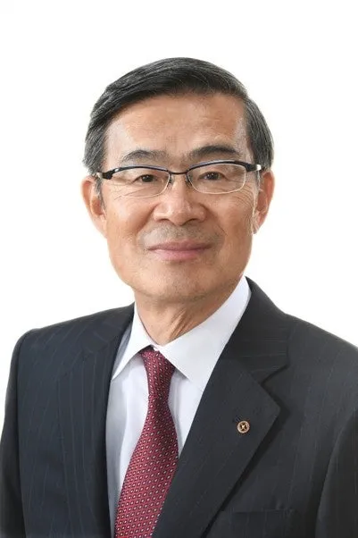 男性が正面を向いて立っているポートレート。彼は眼鏡をかけ、ダークスーツと赤いネクタイを着ています。背景は白です。