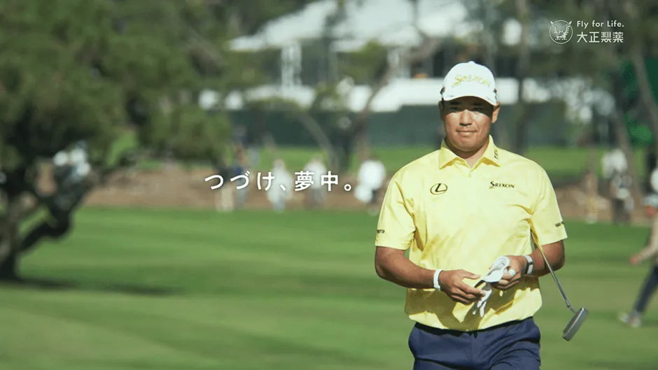 ゴルフコースを歩く男性。彼は黄色のシャツと白い帽子をかぶっています。彼の左側には緑の木があり、背景には他の人々がいます。