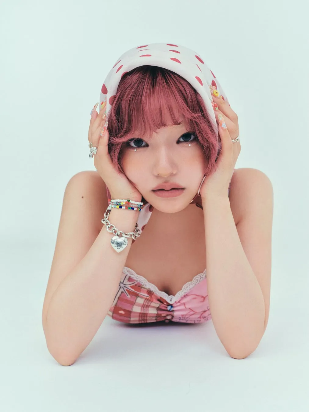 ピンクの髪の女性が顔を両手で支え、カメラ目線でポーズをとっている写真。