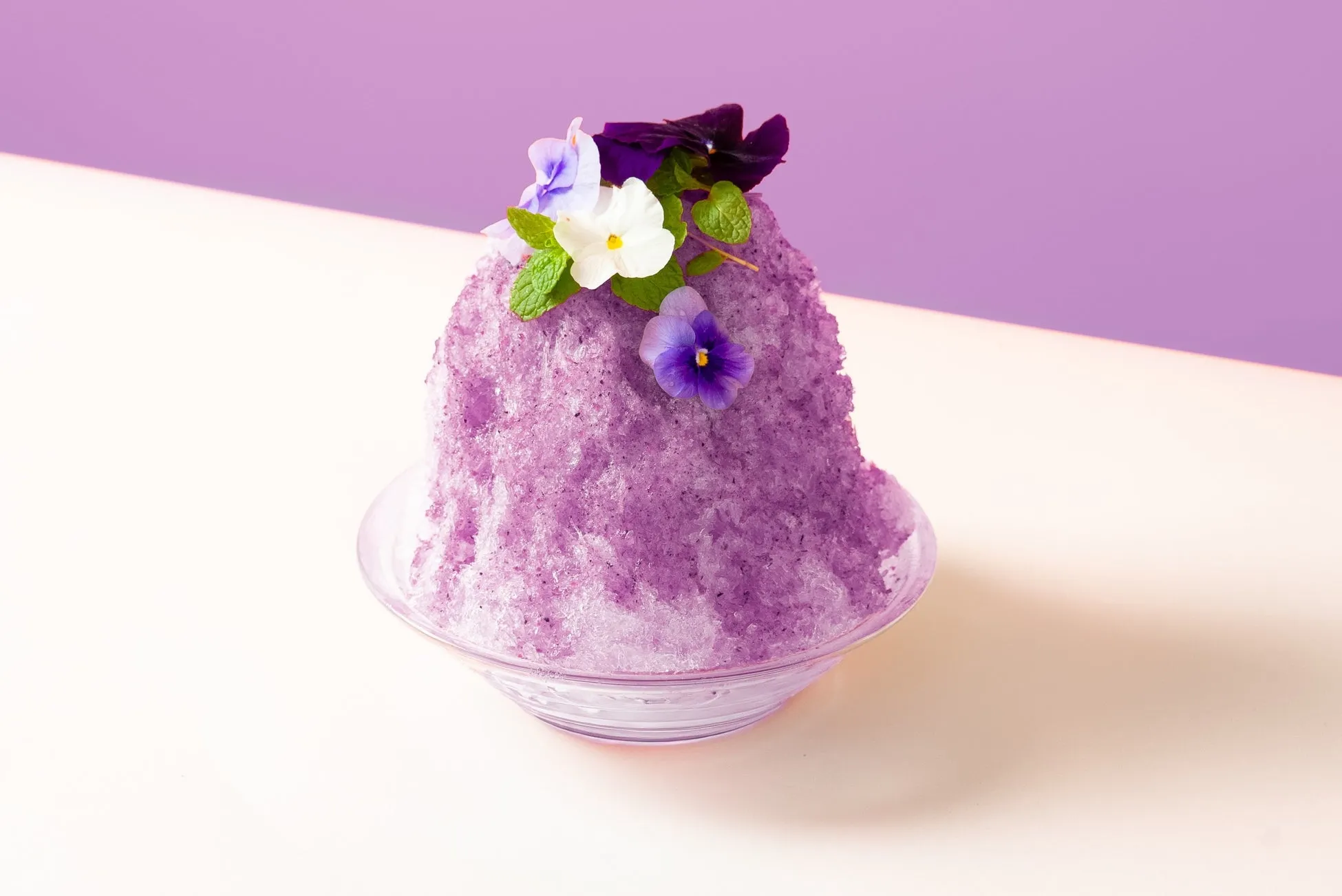 紫色のカキ氷。上に花が飾られている。透明なガラスの器に入っている。