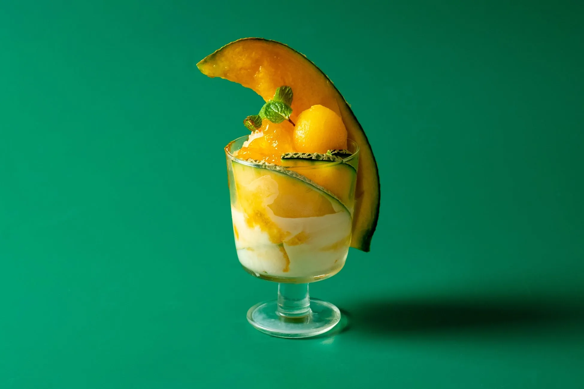 メロンをあしらったデザートのグラス。緑色の背景に、メロンの果肉やミントが添えられており、見た目も美しい。