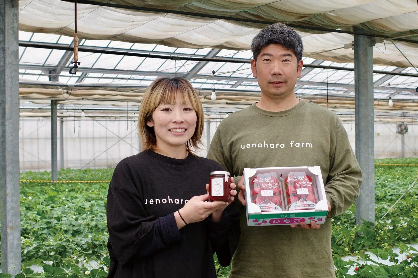 二人の人物がイチゴのジャムとパックを手に持っている写真。温室の中で撮影されており、背後にはイチゴの栽培地が見える。