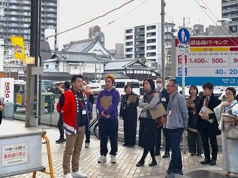 通りで話をする人々。赤い服を着た男性が話をしており、周りには多くの人が見ている。背景には駐車場や建物が見える。