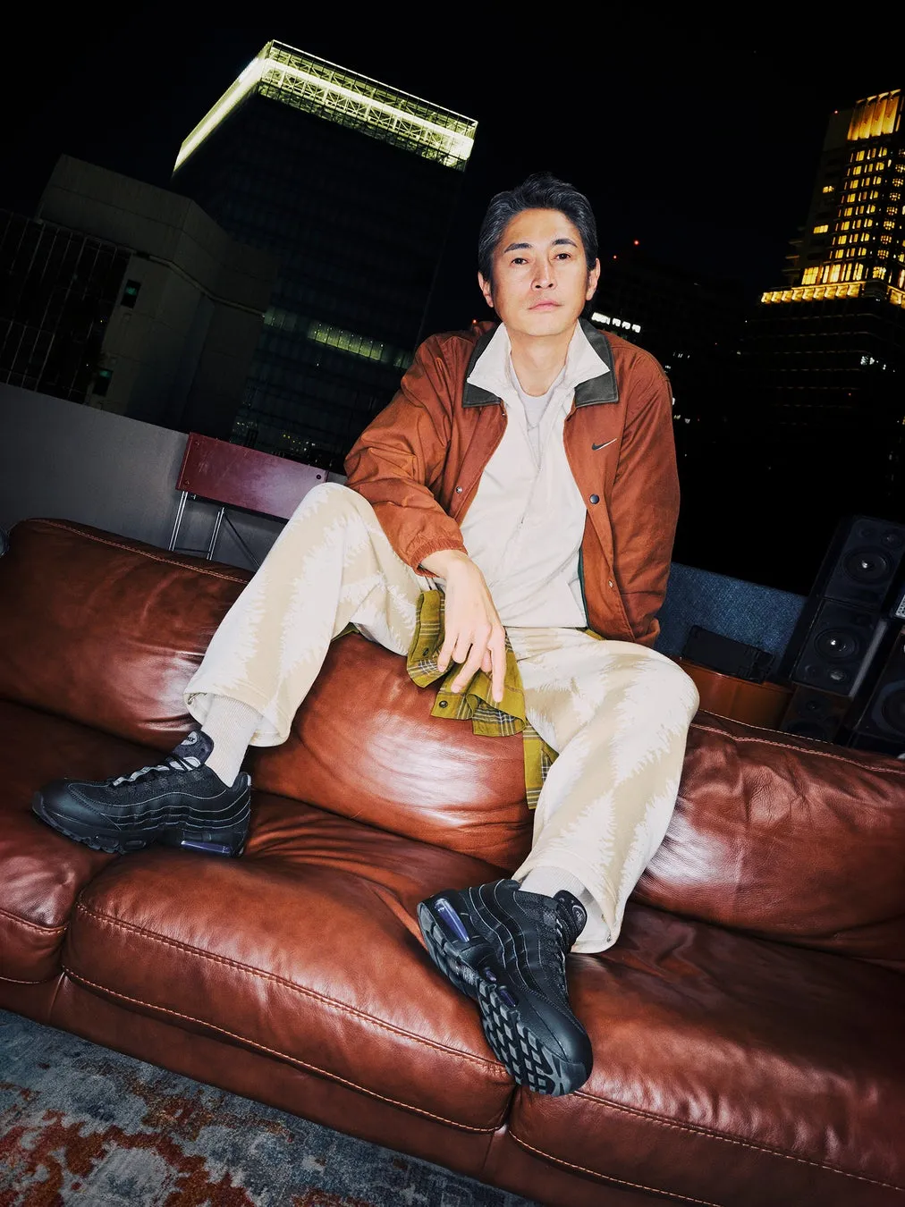 茶色の革張りのソファに座る男性の写真。男性は茶色のジャケット、白いパンツ、黒いスニーカーを履いている。背景には夜の街並みが写っている。