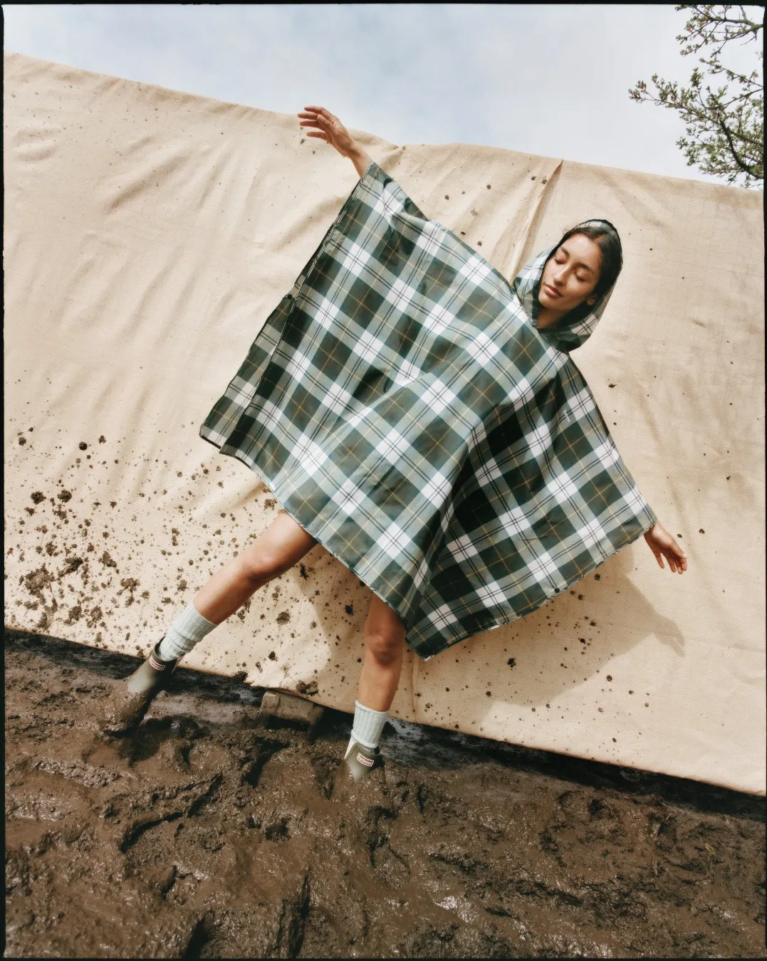 チェック柄のポンチョを着て泥の中に立っている女性の写真です。女性は目を閉じ、腕を広げ、まるで踊っているようです。