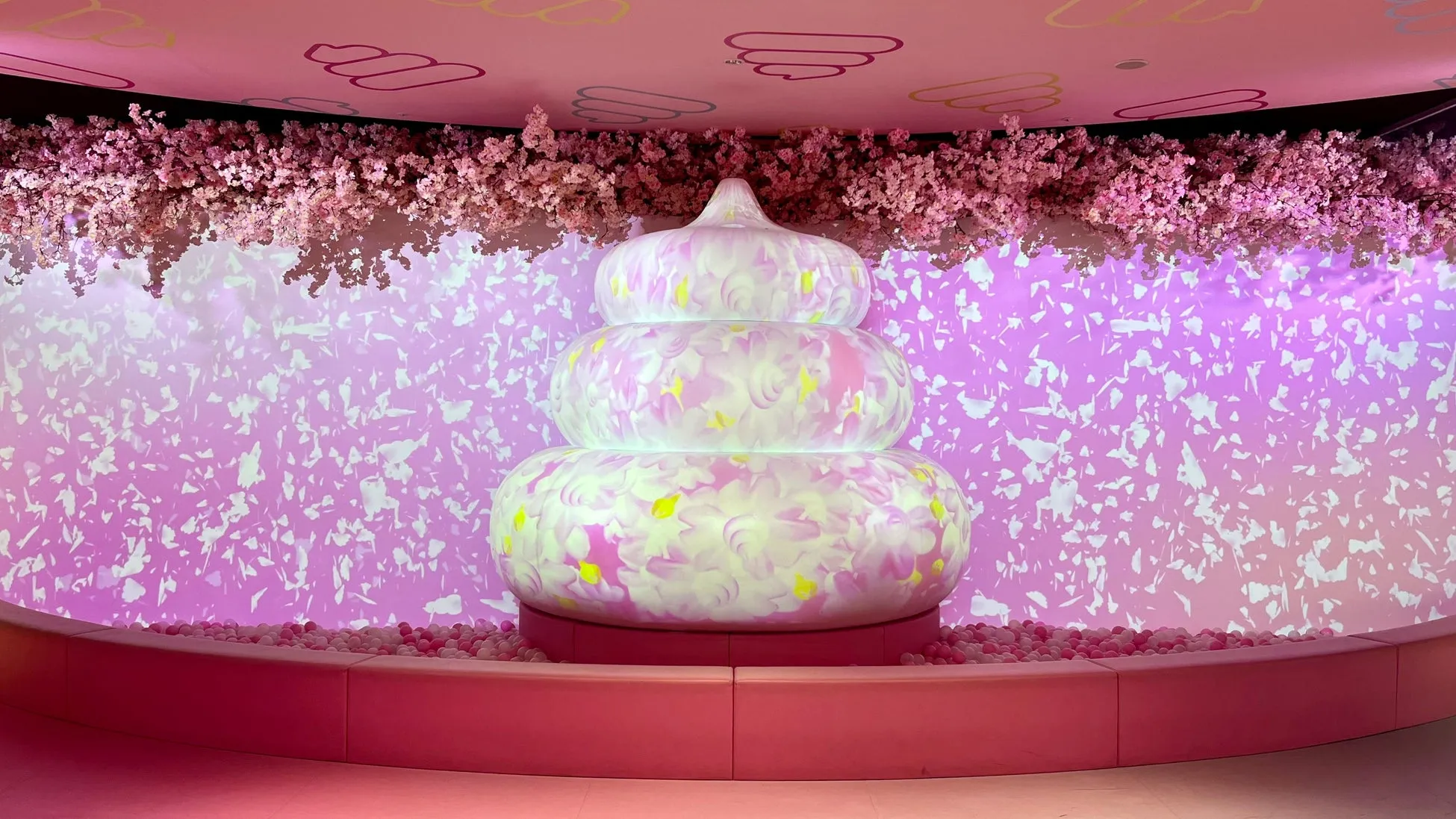 ピンク色の空間に、巨大なオブジェが置かれた写真。オブジェは、ピンクと白の花柄で装飾されており、その周りにはピンクのボールプールが広がっている。壁には花びらの模様が映し出され、天井には花の装飾が施されている。