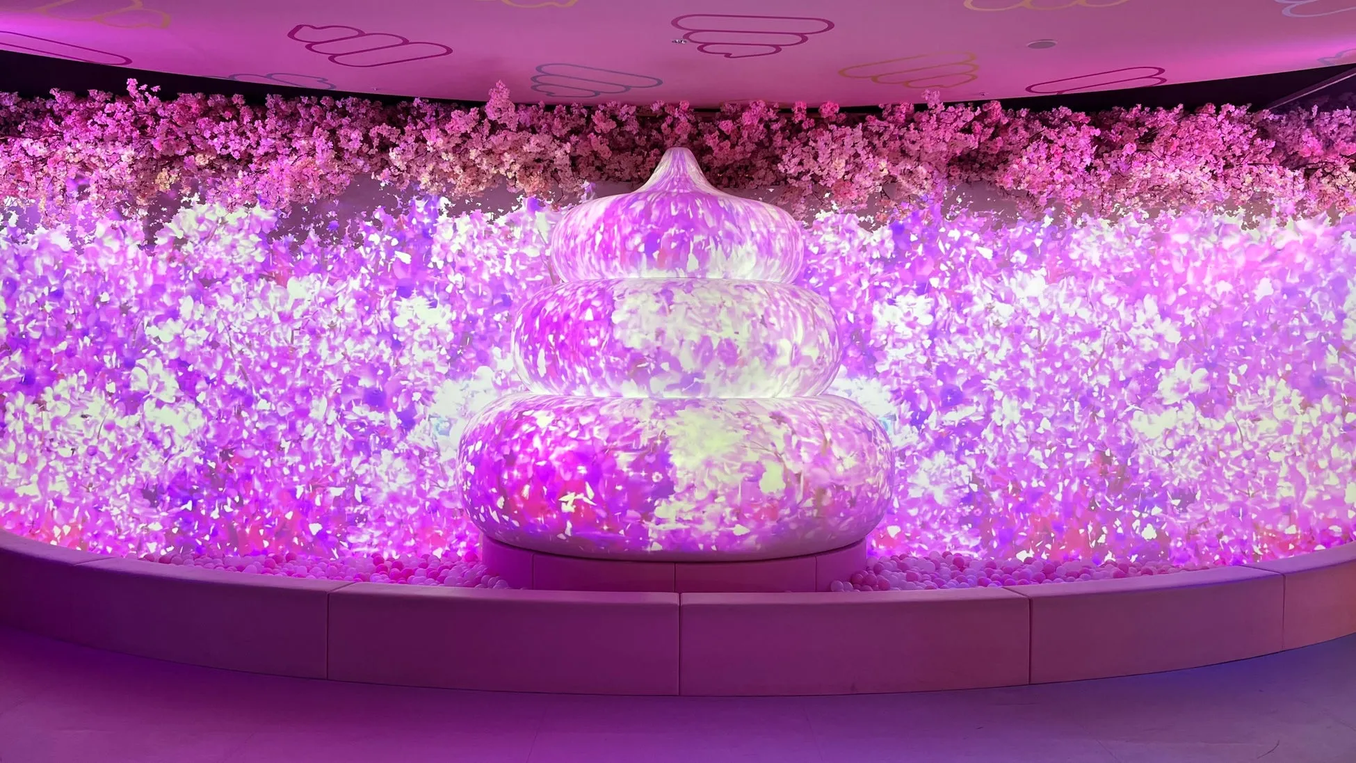 ピンク色の空間に、オブジェと桜のような装飾が施された写真です。オブジェは積み重なったような形をしており、表面には花のような模様が映し出されています。背景には桜の装飾があり、全体的に幻想的な雰囲気です。