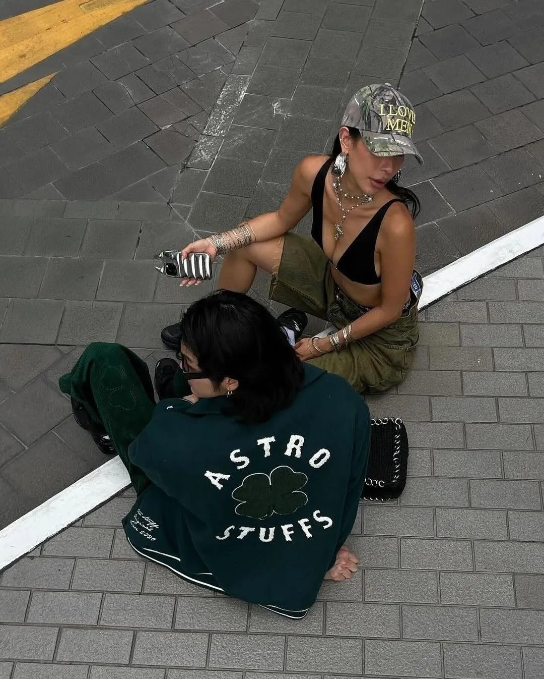 2人の若い女性が、屋外の舗装された場所で座っています。一人はカモフラージュ柄の帽子をかぶり、黒いブラトップとカーキ色のパンツを着ています。もう一人は緑色のジャケットを着ています。二人はアクセサリーを身につけています。