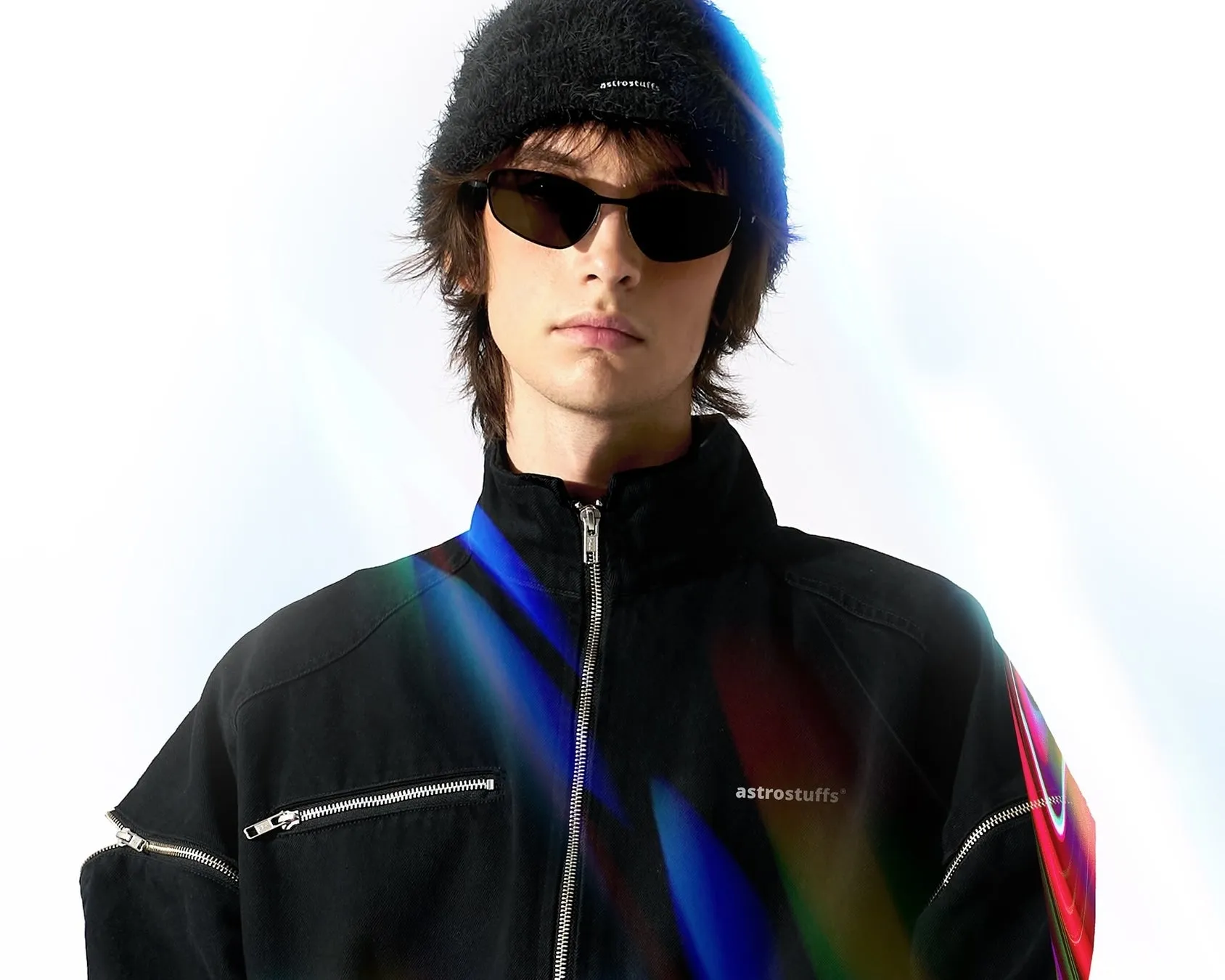 サングラスとニット帽をかぶった男性が、ジッパー付きの黒いジャケットを着て立っています。ジャケットには「astrostuffs」のロゴが入っています。