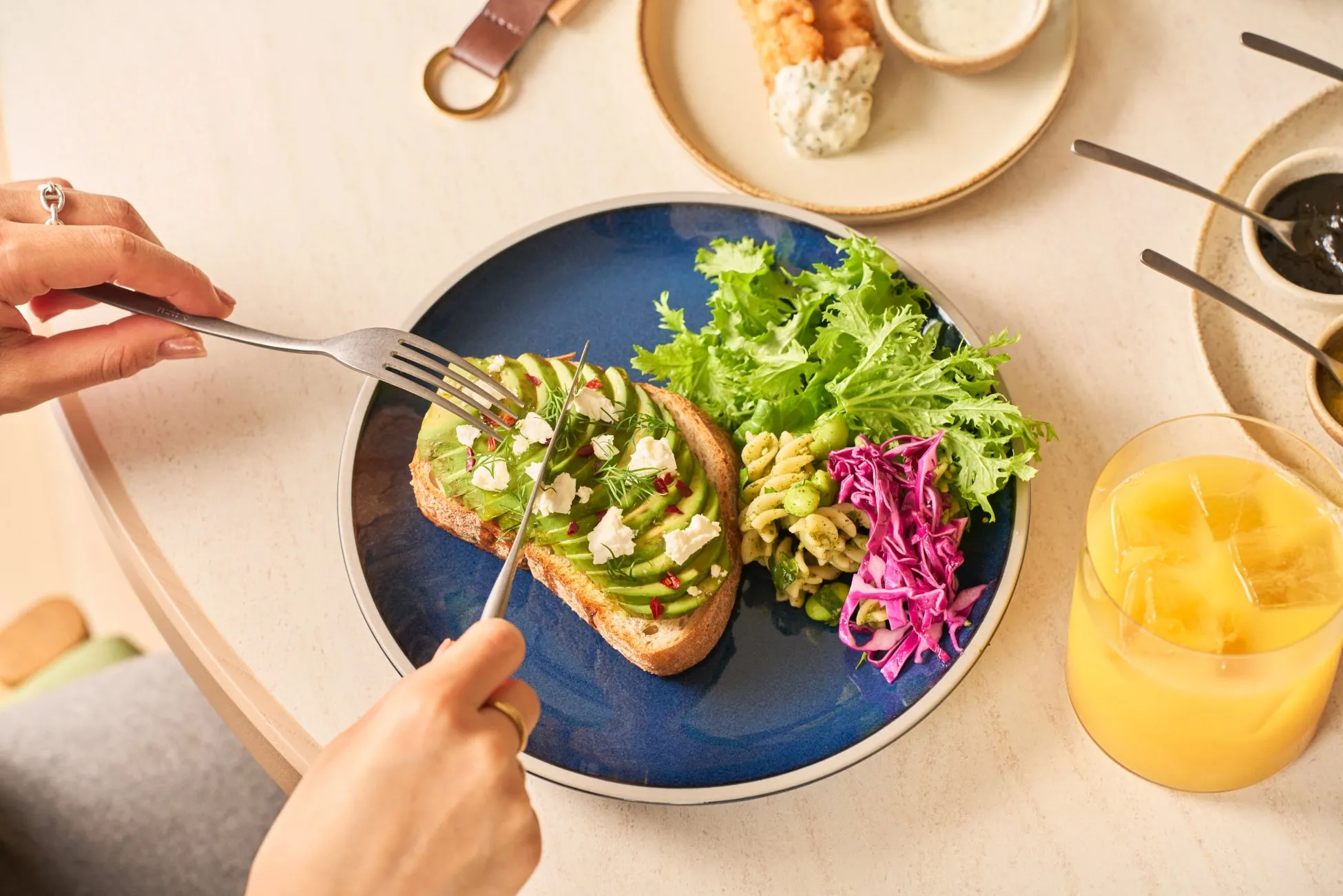 アボカドトースト、サラダ、パスタ、オレンジジュースなどの料理がテーブルに並べられた写真です。フォークでアボカドトーストを食べる様子が写っています。