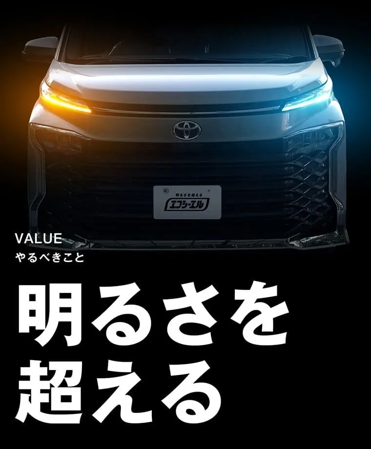 トヨタ車のヘッドライト部分の画像。ヘッドライトはオレンジと青に光っており、車のエンブレムやナンバープレートが見える。背景は黒。