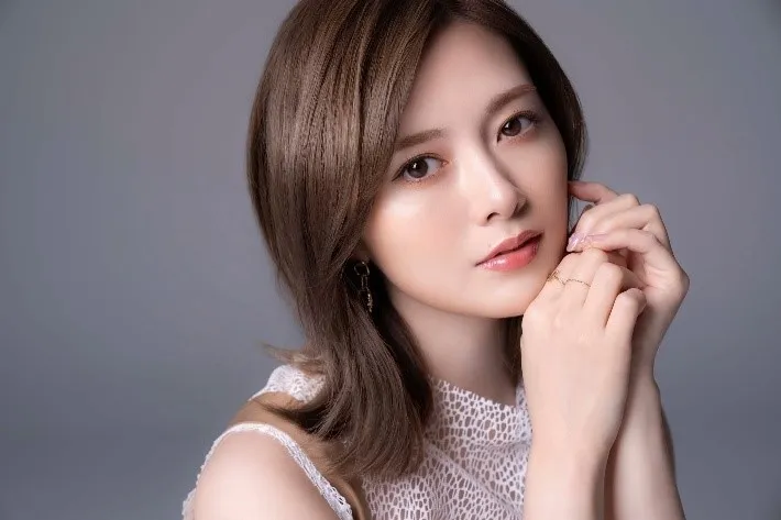 女性のポートレート写真。女性はカメラを見て、顔に手を添えている。彼女は白いレースのトップスを着ている。