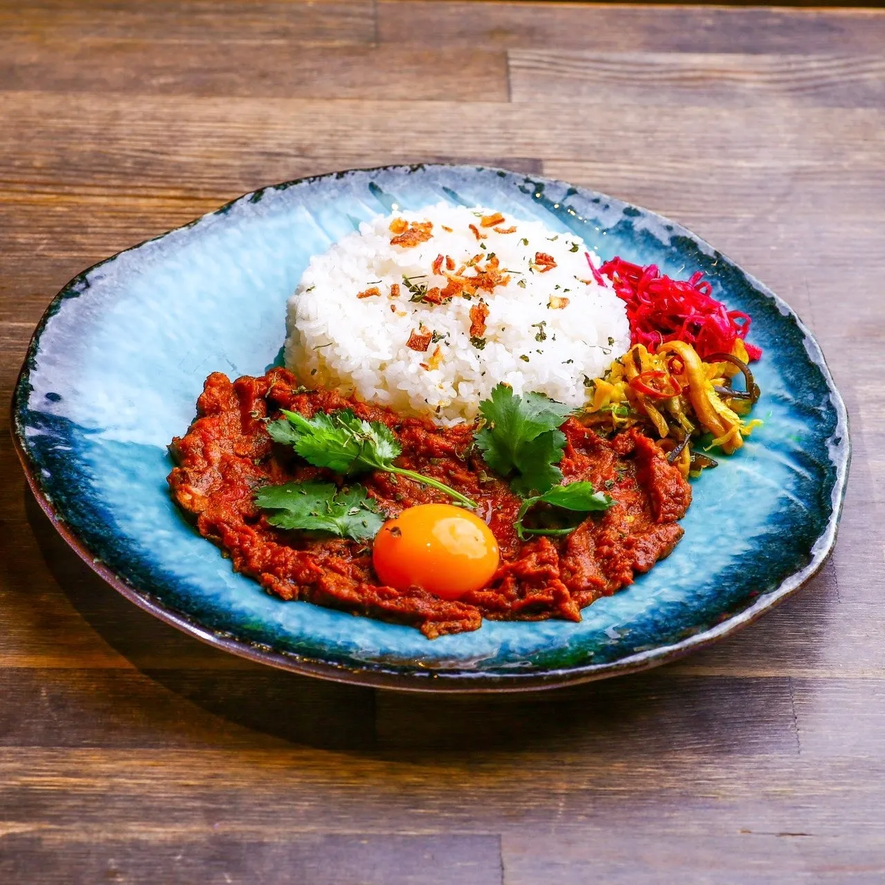 青い皿に盛られた料理の写真。ご飯、赤いソース、卵黄、パクチー、漬物のようなものが写っている。木目のテーブルの上に置かれている。