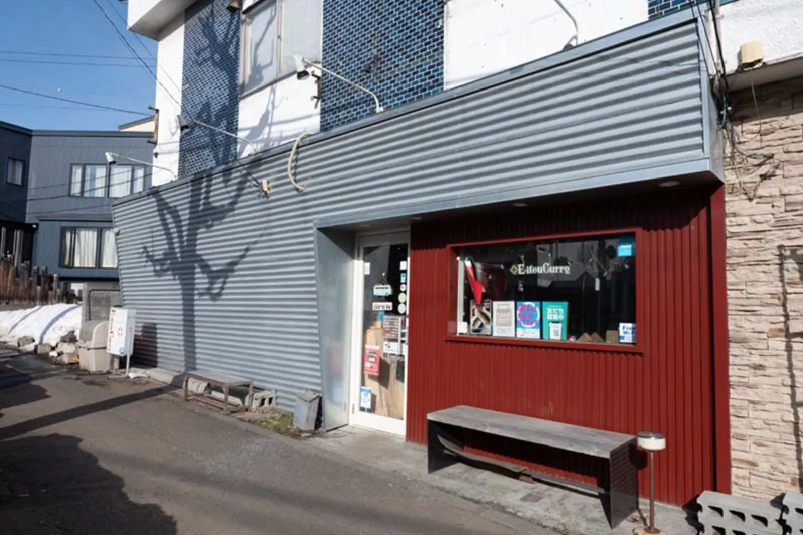 カレー屋さんの外観を写した写真です。赤色の壁に窓とベンチがあり、お店のロゴやメニューが窓に表示されています。