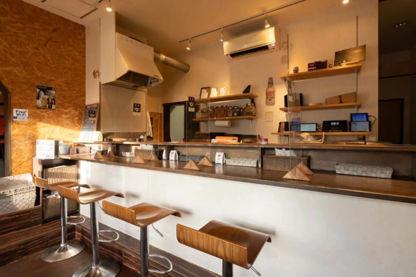 カウンター席のあるカフェの店内。木製の椅子とカウンターが特徴的で、奥には棚があり、様々なものが置かれている。壁にはフリーWi-Fiの案内もある。