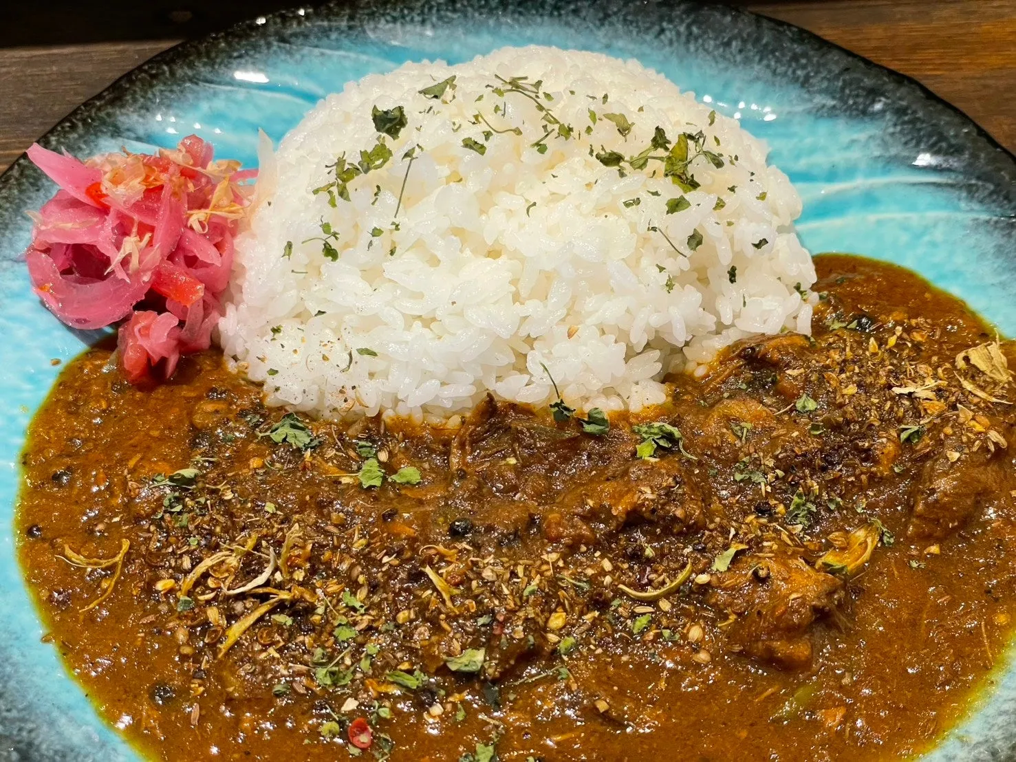 白いご飯の上にカレーがかかった料理の写真。カレーの上にはスパイスが散りばめられ、ピンク色の漬物が添えられている。