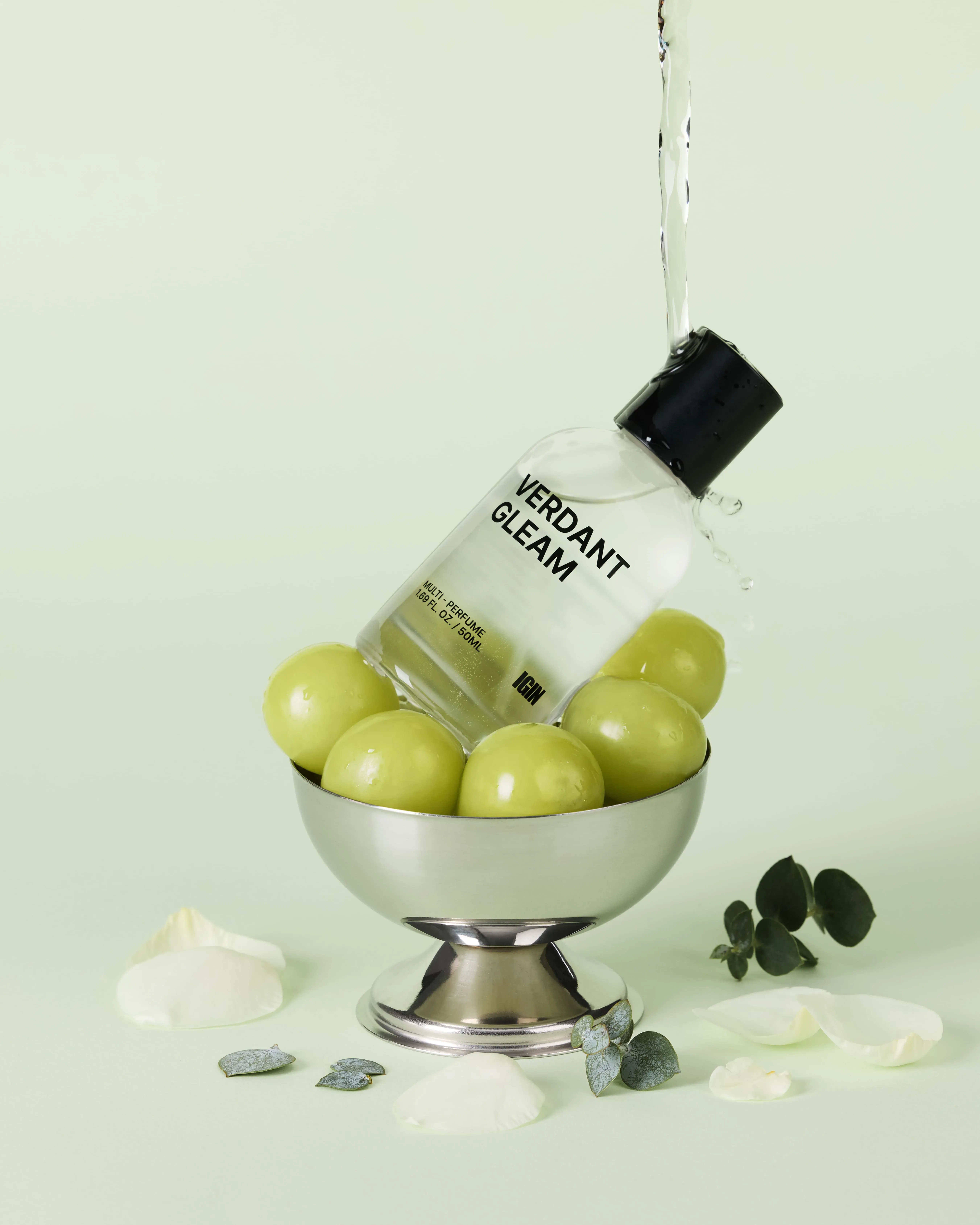 香水ボトルの周りにブドウと花びらを配置した静物写真。香水ボトルからは水が流れ出ている。