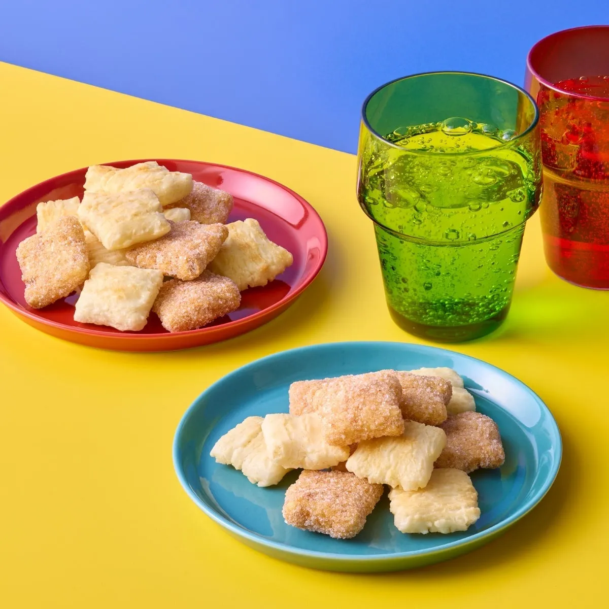 カラフルな背景に、お菓子と飲み物が置かれた写真です。お菓子は、ピンクと青の皿に盛り付けられており、砂糖がまぶされています。飲み物は、緑と赤のグラスに入った炭酸飲料のようです。