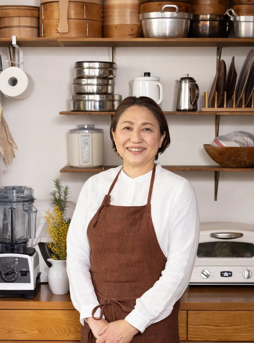 キッチンに立つ女性の写真。彼女はエプロンを着ており、笑顔でカメラを見ている。