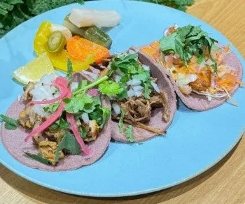 青い皿に盛られた3つのタコスと、ピクルス、オレンジ、レモン、ハラペーニョなどの付け合わせ。