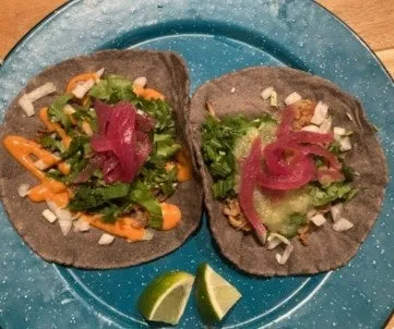 青い皿に2つのタコスとライムのくさびが写っています。タコスは青いトルティーヤで作られており、レタス、紫玉ねぎ、オレンジ色のソースがトッピングされています。