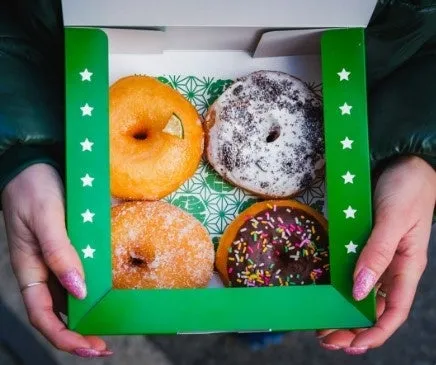 ドーナツの箱を持った人の写真。箱には4つのドーナツが入っており、それぞれ異なるトッピングが施されている。