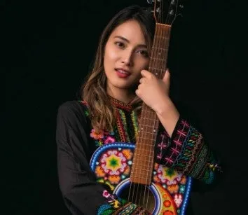 ギターを抱えた女性のポートレート。彼女はカメラ目線で、黒い背景の前に立っています。彼女はカラフルな刺繍が施された黒いシャツを着ています。