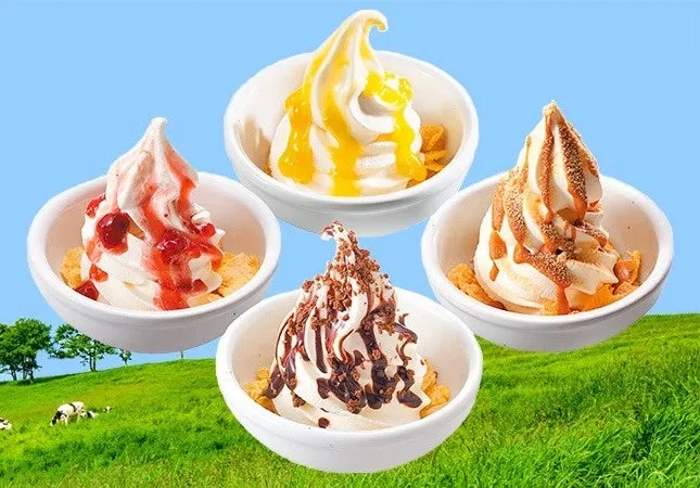 さまざまなトッピングが施されたソフトクリームが4つ、白い器に入っている写真です。背景には、緑の草原と青い空が広がっています。