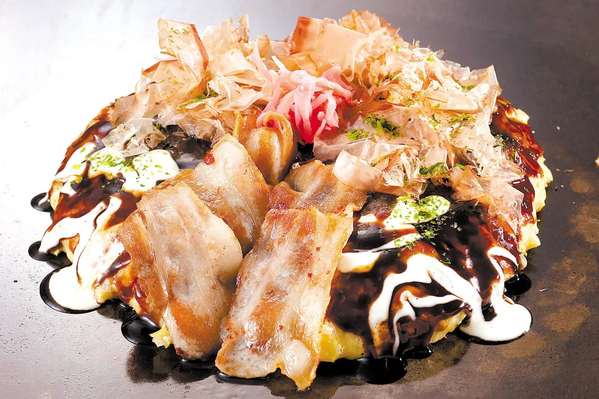 お好み焼きのクローズアップ写真。鰹節、紅しょうが、豚バラ肉がトッピングされている。