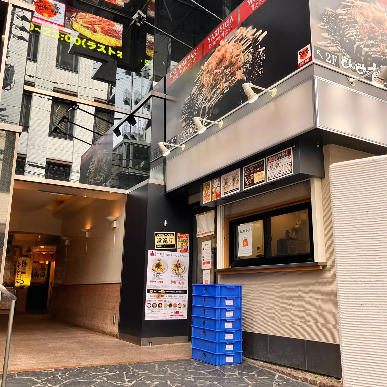 日本のレストランの外観。看板には「OKONOMIYAKI」と「YAKISOBA」の文字があり、テイクアウトの窓や営業中の看板が見える。青い収納ボックスが積み重ねられている。