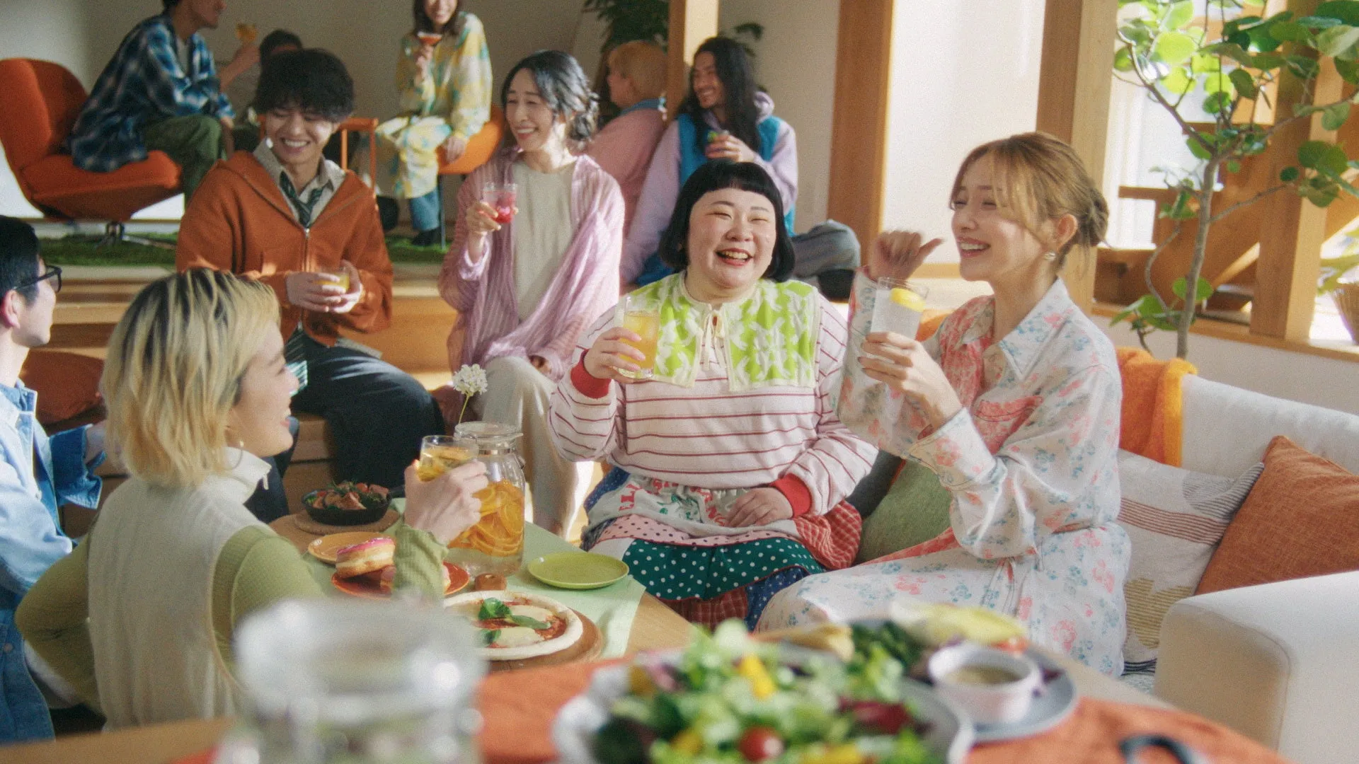 リビングルームで集まる人々の写真。人々は、飲み物、ピザ、サラダなどの食べ物や飲み物を楽しんでいるように見える。陽気な雰囲気があり、リラックスした社交的な集まりのようだ。