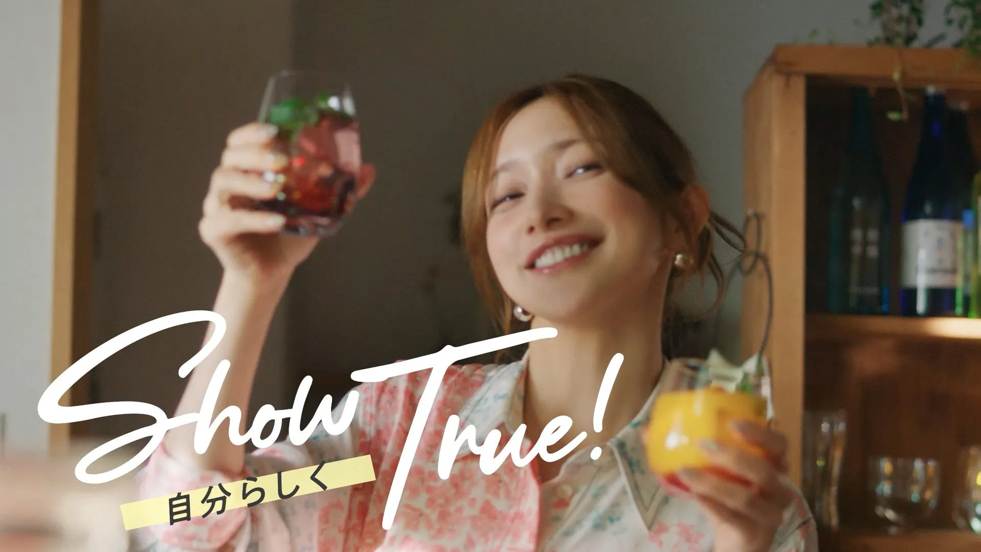女性がグラスを持って笑顔でいる様子。背景には棚があり、飲み物が入ったグラスがいくつか置かれています。