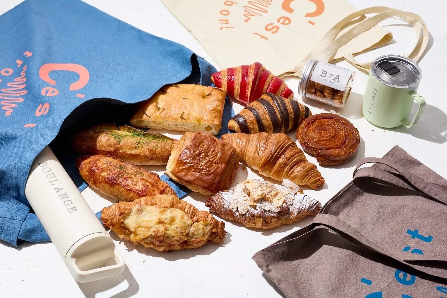 様々な種類のパンが入った青いトートバッグと、パンの入った袋、マグカップ、水筒などが置かれている様子。