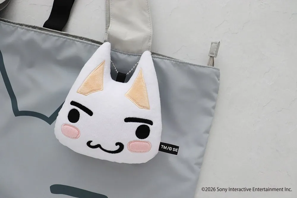 白い猫のぬいぐるみとグレーのバッグが写った写真。猫のぬいぐるみは、黒い目、眉毛、口、そして頬にピンク色のアクセントが施されており、バッグにぶら下がっている。バッグには黒い持ち手が付いており、背景は白い壁。