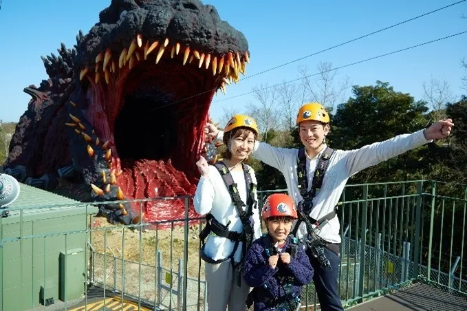 ゴジラの口のようなアトラクションの前に、ヘルメットとハーネスを装着した家族3人が立っている写真です。