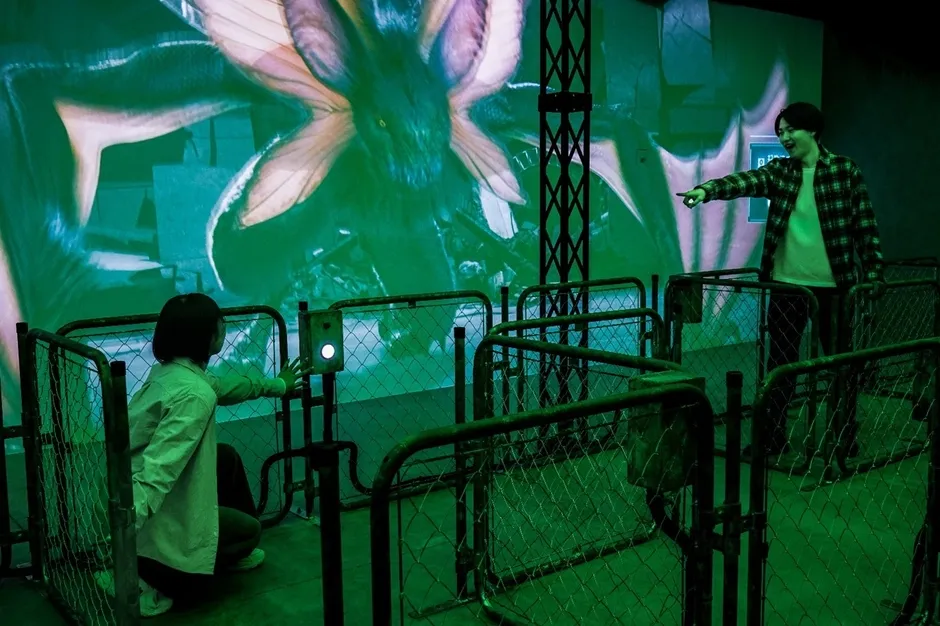 巨大なモンスターの映像が映し出された部屋で、人々がボタンを押したり、映像を鑑賞している様子。