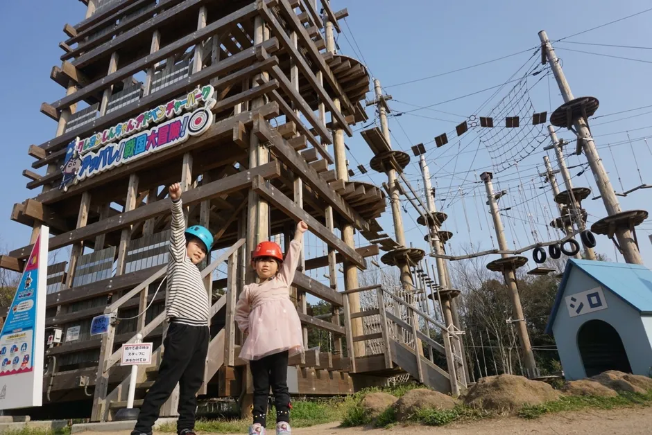 2人の子供がアスレチック施設の前でポーズをとっている写真です。背景には、木製の構造物とロープウェイがあり、青い空が広がっています。