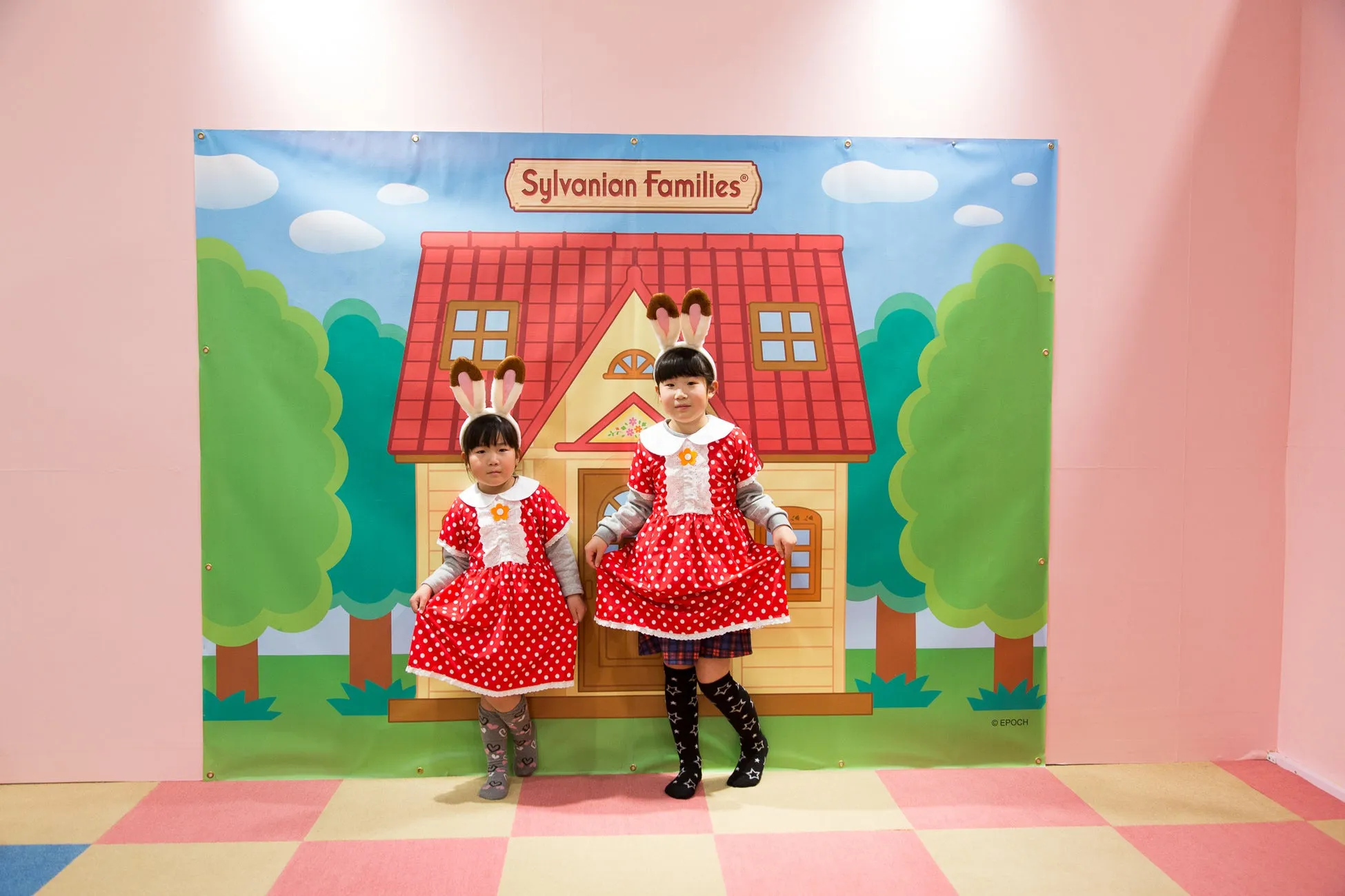 2人の女の子が、赤い水玉模様のドレスとウサギの耳のヘアバンドを着て、シルバニアファミリーの背景の前でポーズをとっています。