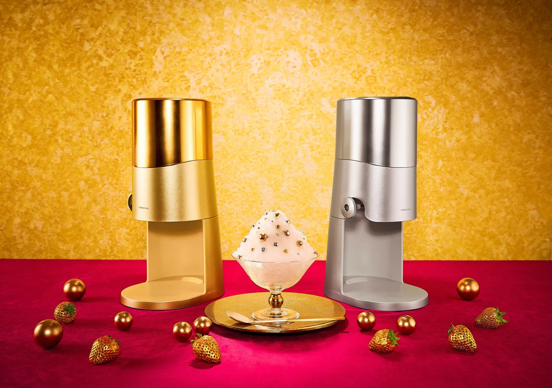 金と銀のかき氷機が並び、ガラスの器に入ったかき氷が中央に置かれている写真。周りには金色のボールと金色のイチゴが散りばめられている。背景は金色の壁。