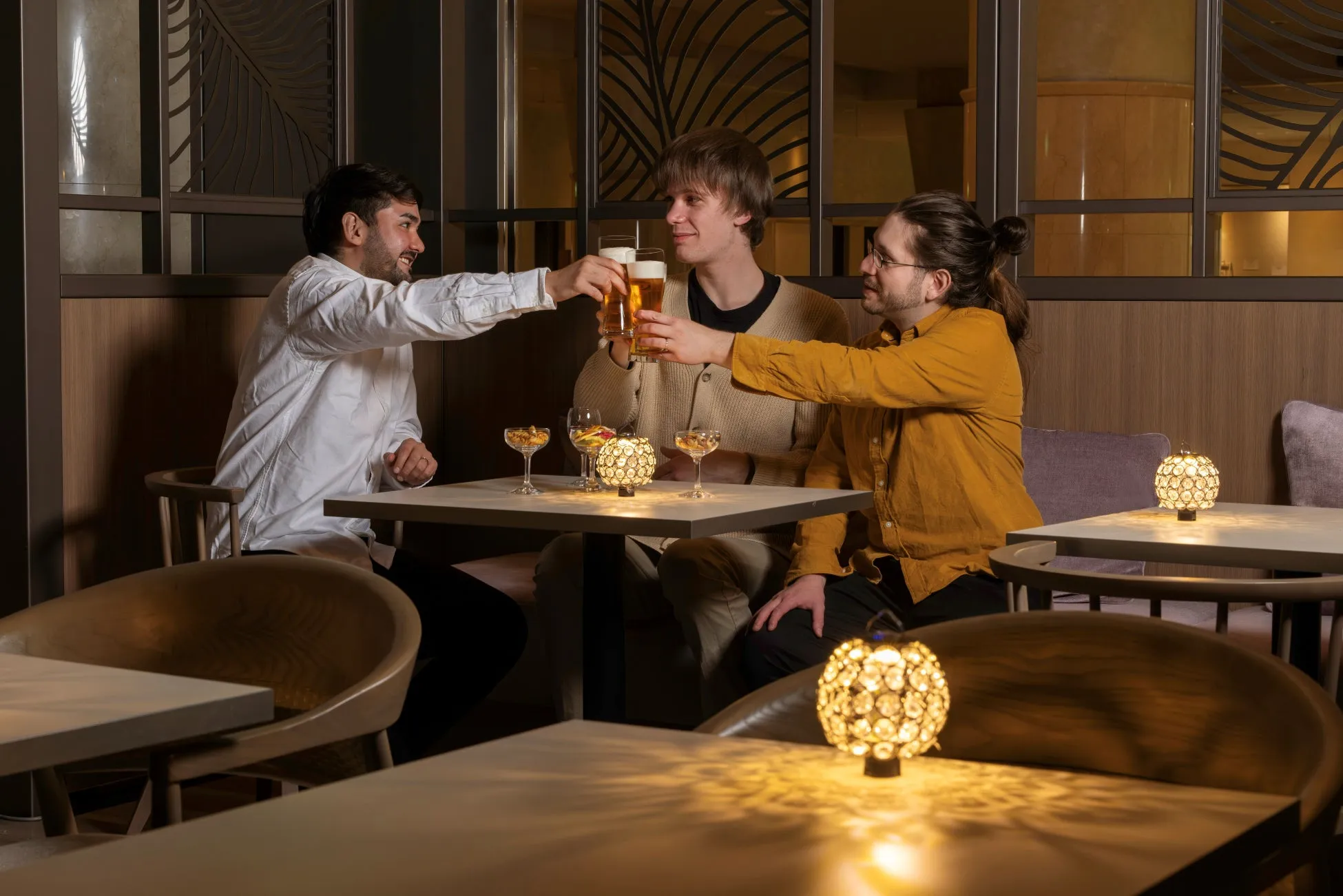 3人の男性がバーのような場所でビールグラスを高く掲げて乾杯している様子を捉えた写真。テーブルには、シャンパングラス、スナック、装飾的なランプが置かれ、親密な雰囲気を醸し出しています。